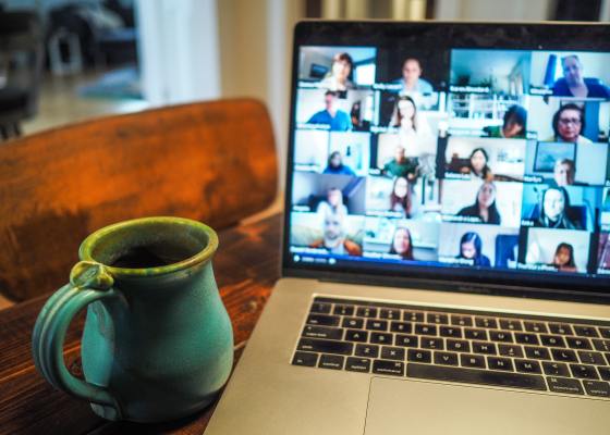 Open laptop with approximately 20 people tiled on the screen in an online meeting. A mug is on the table to the left of the laptop.