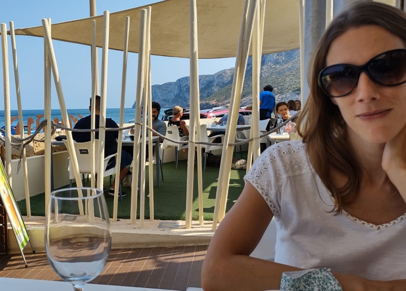 Photo of young woman wearing sunglasses sat at a table outside a restaurant. Sea view behind her and a wine glass on the table.
