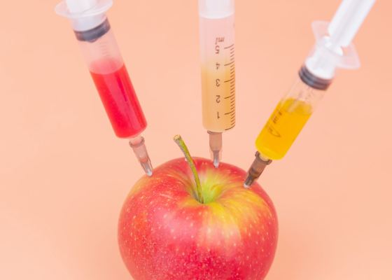 Syringes with different chemicals injections into a red apple on an orange background.