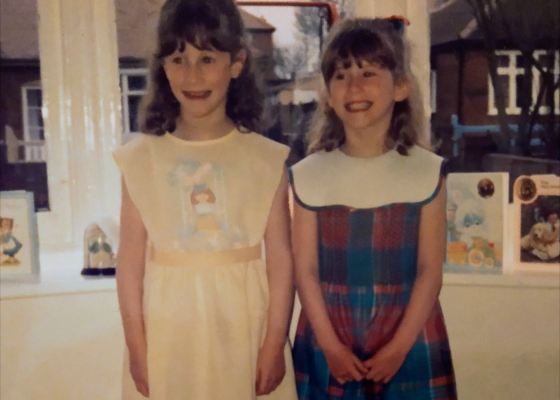 Carly and her sister when they were around 6 years and 5 years old. Carly is wearing a yellow dress and here sis is wearing a blue and pink dress with a white collar. They are both smiling.